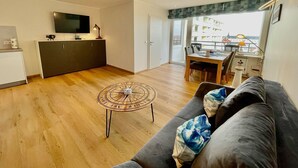 Un espace de vie moderne doté d’un plancher en bois, d’un canapé avec des coussins, d’une table à manger, d’un coin cuisine avec un téléviseur et d’un balcon offrant une vue sur les immeubles.