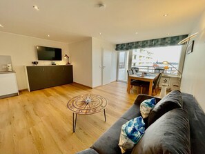 Un espacio habitable moderno con suelo de madera, un sofá con cojines, una mesa de comedor, una zona de cocina con televisor y un balcón con vistas a edificios.