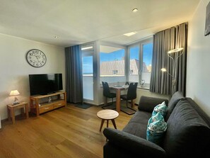 A modern living room with a flat-screen TV, a sofa, a dining table with chairs, and a balcony with a view. - Strandblick im Haus Seeblick Bismarckstraße 6. Etage - Seeblick (Borkum)