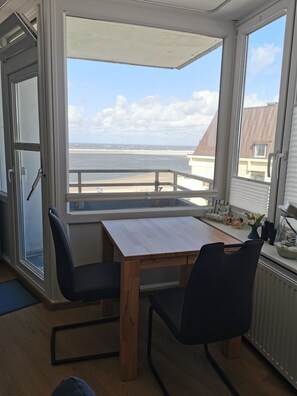 A room with a wooden table and two chairs, a view of the beach, and a balcony with a railing.