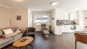 A modern living room with a sofa, armchairs, a coffee table, and a kitchen area with cabinets and appliances. - Sommerwind Alfonso Borkum Apartment 1 - Vacation with dog - Sommerwind Alfonso (Borkum)