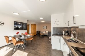 A modern kitchen with a built-in oven, a sink, and a coffee maker. There is a dining area with a table and chairs, and a living area with a sofa and a TV mounted on the wall.