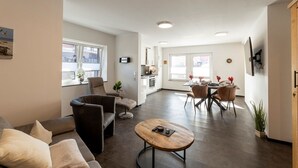 A modern living room with a sofa, armchair, and coffee table, a dining area with chairs and a table, and a kitchen in the background. - Sommerwind Alfonso Borkum Apartment 1 - Vacation with dog - Sommerwind Alfonso (Borkum)