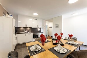 Una cocina moderna con armarios blancos, encimera negra y comedor con una mesa de madera preparada para cuatro comensales.