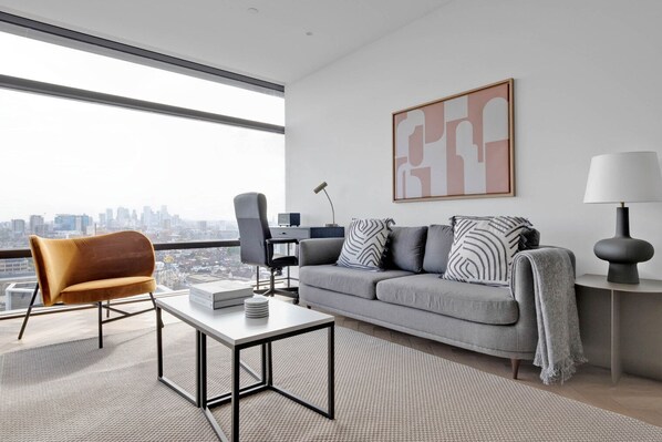 Un salon moderne avec un canapé, un fauteuil et une table basse, une vue sur la ville et un tableau abstrait encadré.