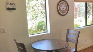 Una habitación con una mesa redonda de madera y dos sillas, ventanas amplias con persianas, una lámpara de pared y un cartel de bienvenida.