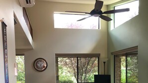 Un dormitorio con cama, ventilador de techo, televisor y ventanas amplias que dan a vista de árboles.