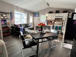 A modern kitchen with white cabinets, a dining table with chairs, and a living area with a sofa and a TV. - Artful Cottage Mtn. Retreat - 55 or over (Waynesville)