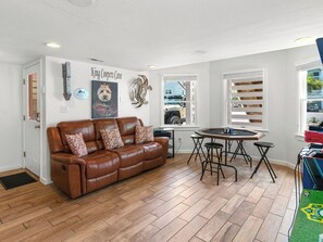 A living room with a brown leather sofa, a small round table with stools, a wall-mounted artwork, and a window with blinds. - The Silly Geese: Pool Hot Tub Games, Walk to Beach (Corolla)