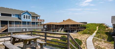 Een houten terras met picknicktafels, een zandpad en gebouwen aan het strand.