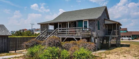 A wooden house with a porch and stairs, surrounded by sand and vegetation.