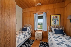 A wooden-paneled room with two beds, a small table, and a window with curtains. - Oceanfront, dog friendly, walkway to beach, cozy historic cottage- Island Dancer (Nags Head)