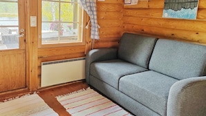 A cozy cabin interior with a grey sofa, wooden walls, a window with a view of greenery, and a door with a curtain. - Vacation home Ainola by Interhome (Joutsa)