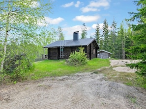 A rustic log cabin with a chimney, surrounded by a dirt path and dense forest.