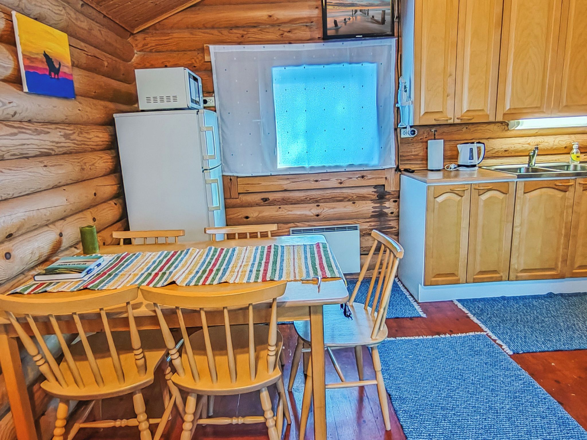A cozy cabin-style kitchen with wooden walls, a dining table with chairs, a refrigerator, a microwave, and a window.
