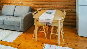 Un interior de cabaña acogedor, con una pared de madera, un sofá gris, una mesa de comedor de madera con sillas y un refrigerador blanco.