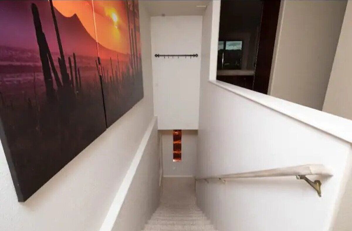 Una escalera estrecha con barandilla blanca, un pequeño ventanal en la parte superior y un perchero adherido a la pared.