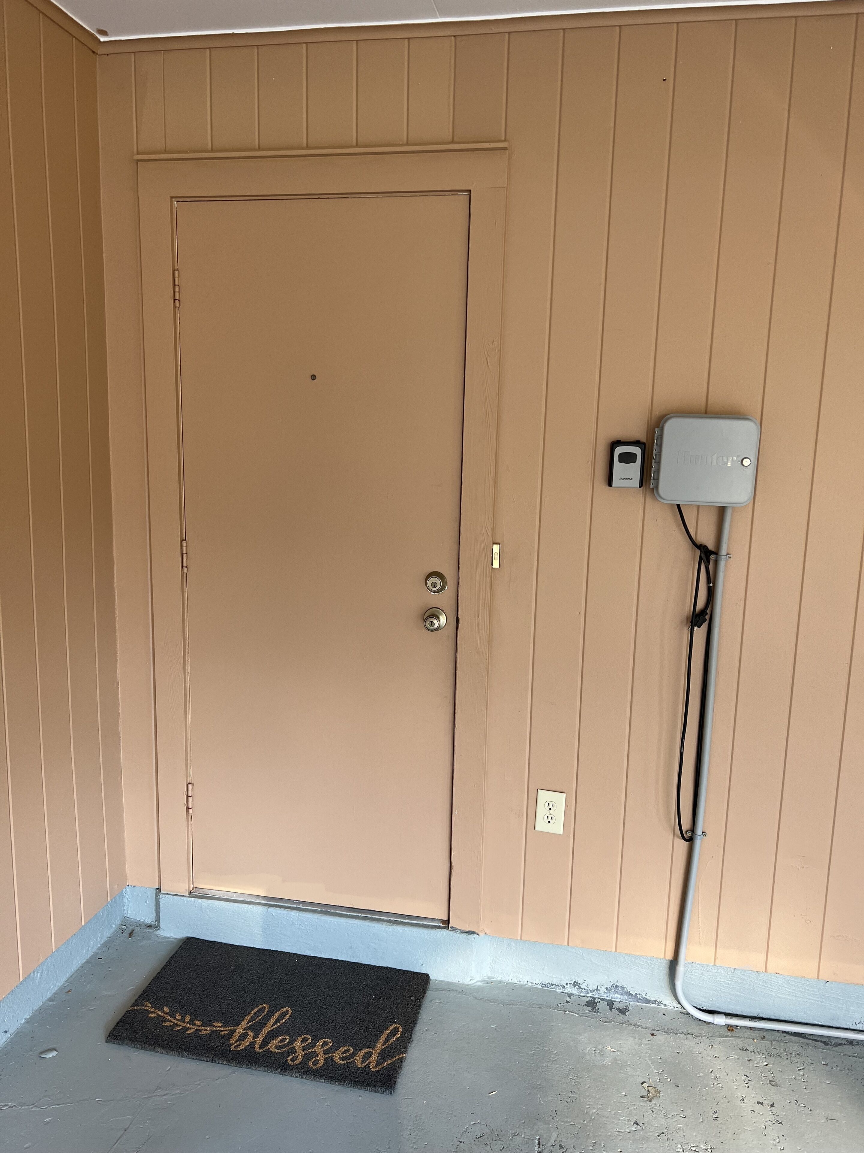 Una puerta color beige con picaporte y una pequeña caja eléctrica fijada en la pared a su lado.