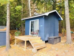 Situated in a wooded area, this cabin features a front door with a glass panel, a small porch with a bench and table, and wooden steps leading up to the entrance.