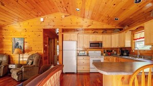 A cabin interior with a wooden ceiling, a kitchen with wooden cabinets, a refrigerator, a microwave, a stove, and a dining area with a bench and a chair.