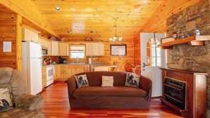 A log cabin interior with a wooden ceiling, stone wall, kitchen, sofa, fireplace, and wooden floors.