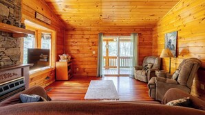 A log cabin interior with a stone fireplace, wooden floors, a ceiling fan, and a view through a glass door to the outside.