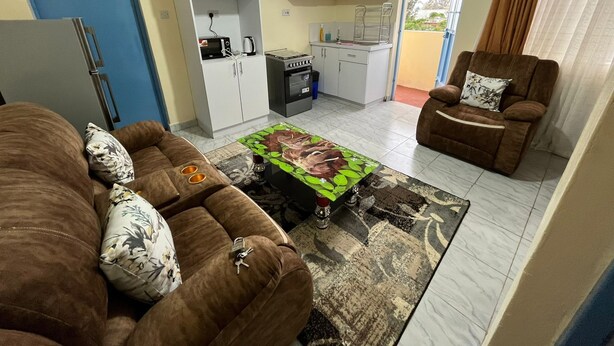 A living room with a brown sofa, a coffee table with a floral design, and a brown armchair with a cushion. There is a kitchen area with white cabinets and a stove.
