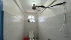 A small, narrow bathroom with white marble walls, a white toilet, a red trash bin, a window, and a shower head.