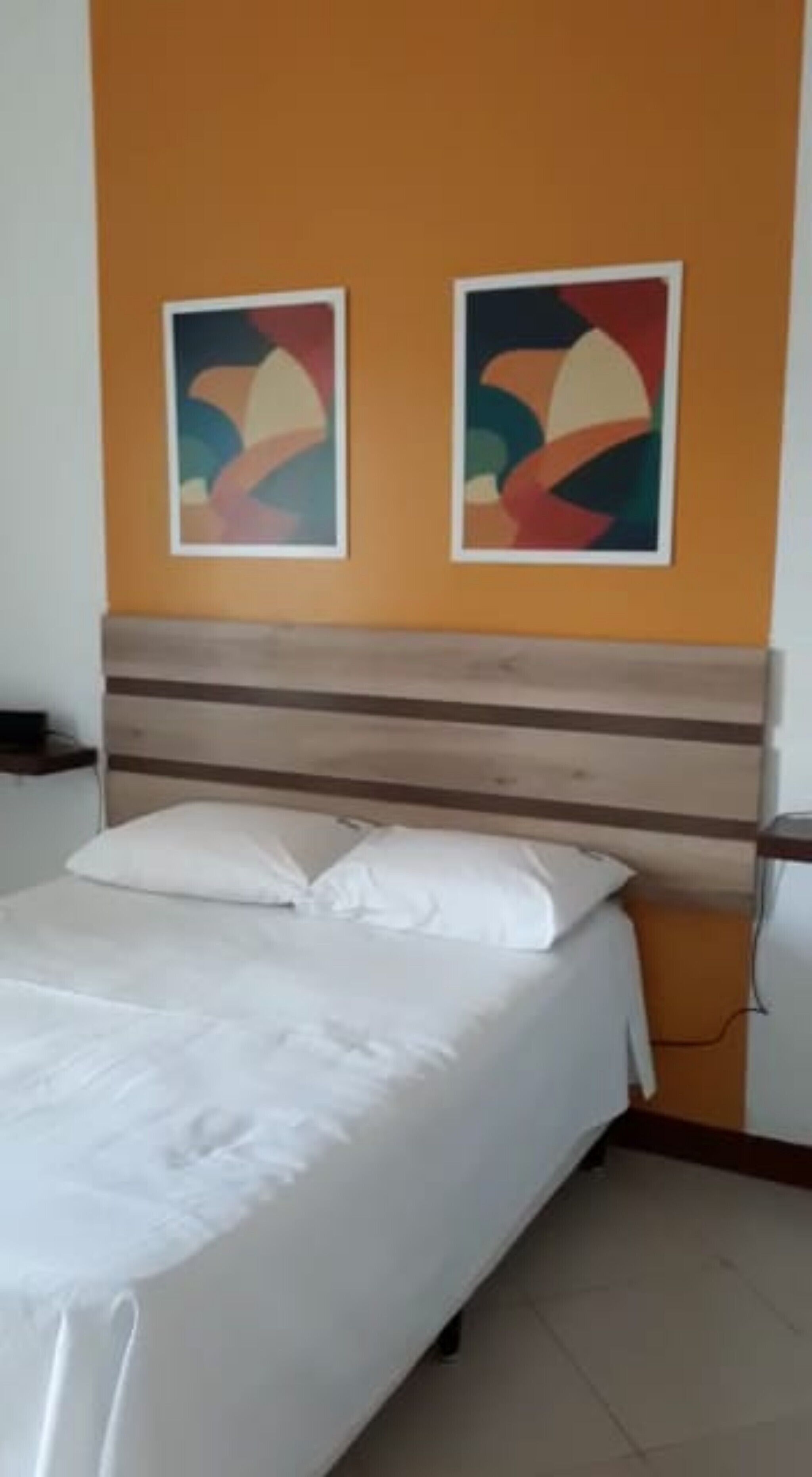 Cama bem arrumada com lençóis brancos num quarto com cabeceira de madeira e duas pinturas abstratas na parede laranja.
