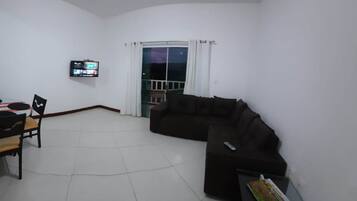 Una sala de estar con un sofá negro, un televisor montado en la pared, una mesa de comedor con sillas y un balcón con cortinas blancas.