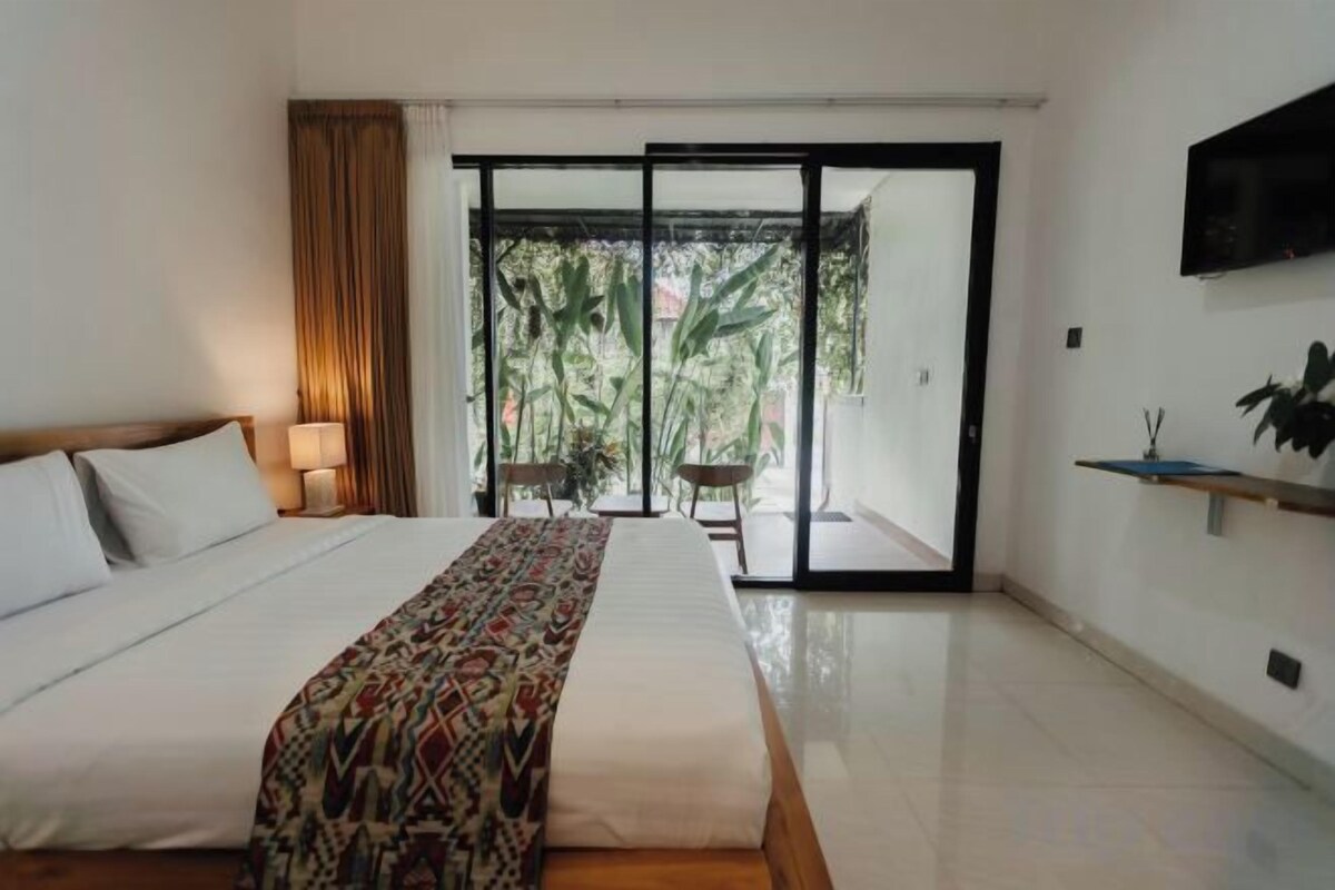 Una habitación de hotel con una cama grande, un ventanal corredero que da a un balcón y un televisor montado en la pared.