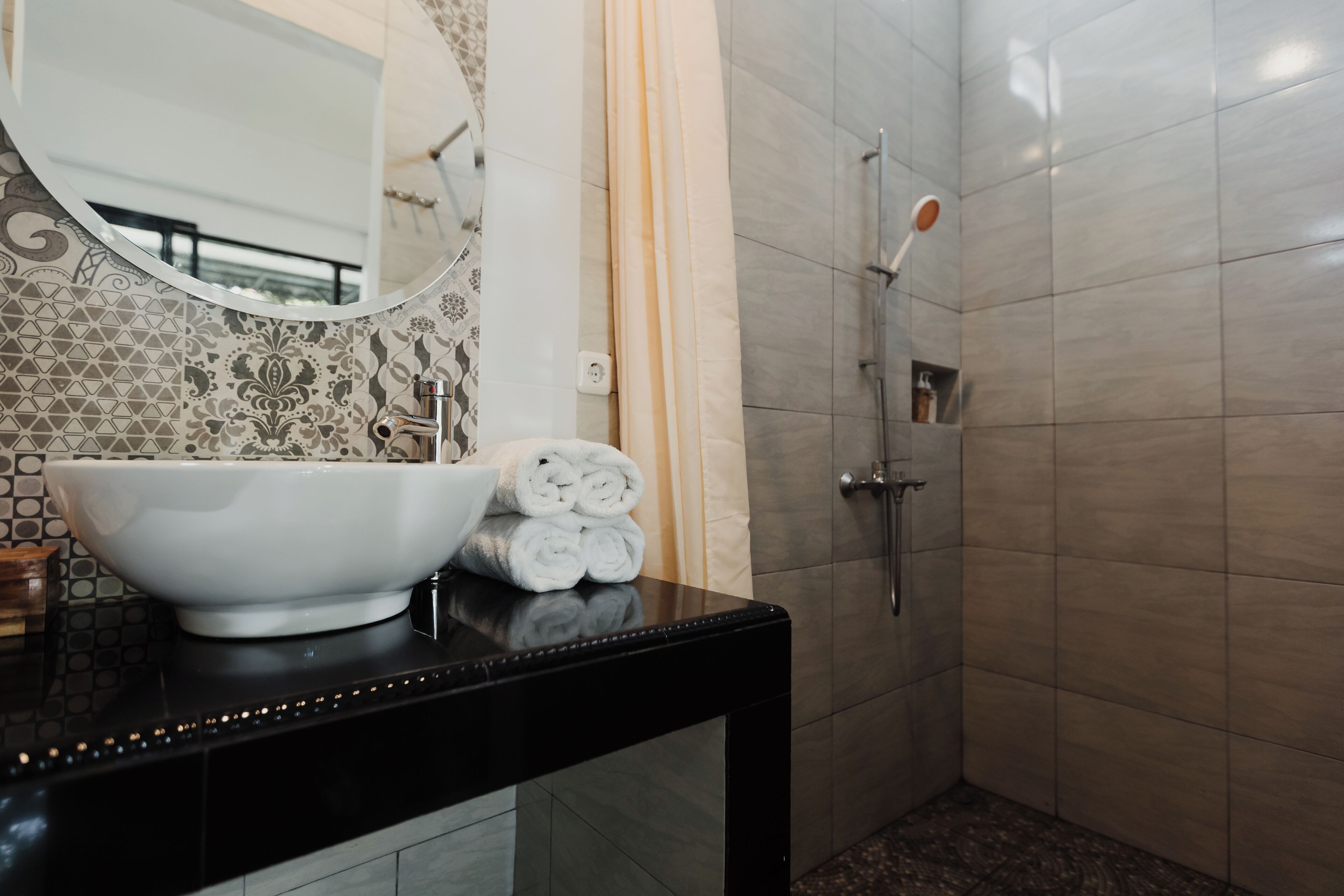 Een badkamer met een zwart aanrecht, een witte wastafel, een spiegel, een douche met handdouche en een handdoekrek met opgevouwen handdoeken.