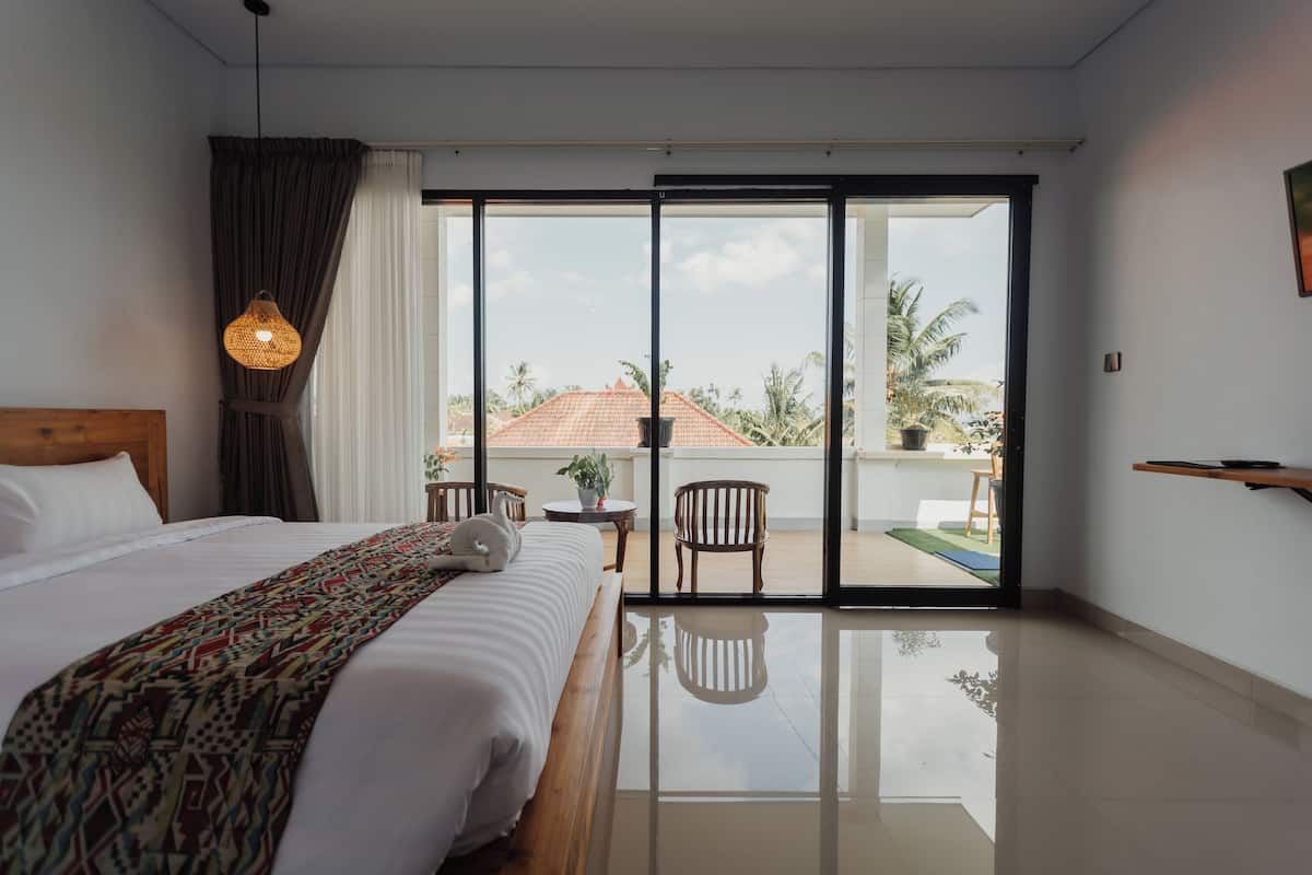 A hotel room with a large bed, a balcony with chairs, a small table, and a view of palm trees.