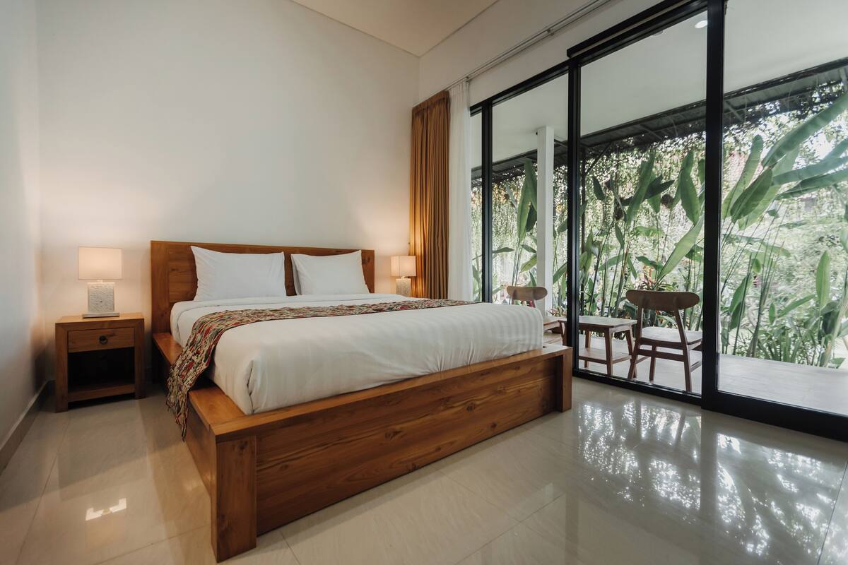A modern bedroom with a wooden bed, two bedside tables with lamps, a large window with a view of greenery, and a small balcony with a table and chairs.