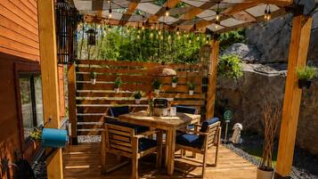 An outdoor wooden pergola with a table and chairs, surrounded by greenery and rocks.