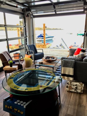 A cozy room with a glass table featuring a Pac-Man game, comfortable seating, and a view of boats on the water. - Seacliff- Luxury Halifax Waterfront Paradise -Pool, Hot Tub, Wharf & Boathouse (Halifax)
