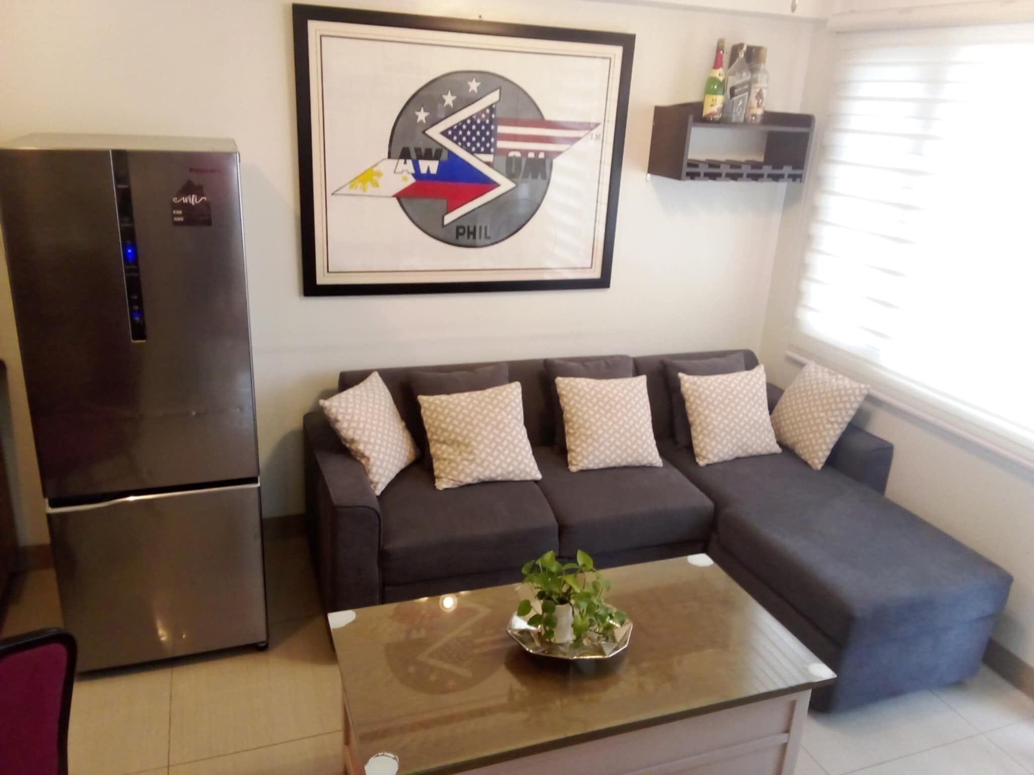 Una sala de estar con un sofá seccional, una mesa de centro con una planta, un refrigerador y un cuadro en la pared.
