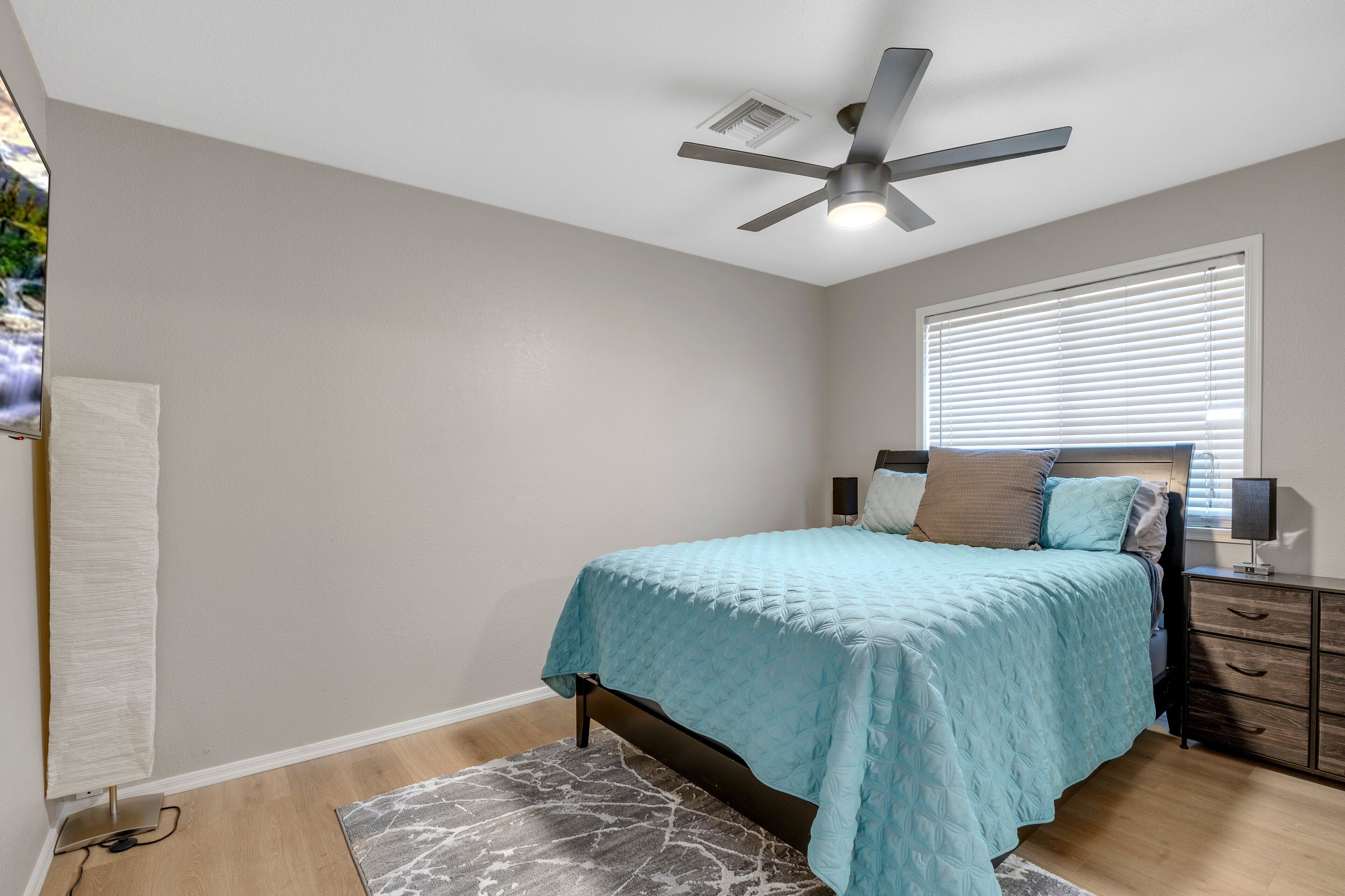 Une chambre à coucher comprenant un lit, un ventilateur de plafond, une fenêtre avec des stores, une table de chevet avec une lampe et un tapis.
