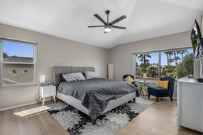 Un dormitorio con cama, mesita de noche, lámpara, silla y vista a palmeras.