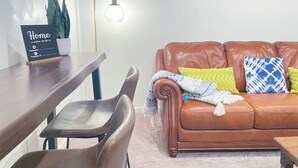 Una sala de estar con un sofá de cuero marrón, una mesa de comedor con sillas, una lámpara de pared y tapices decorativos de tejido.