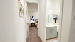Un pasillo que conduce a un dormitorio y un baño. El pasillo tiene piso alfombrado y paredes blancas. Hay un cuadro en la pared. El dormitorio tiene una cama con colcha blanca y almohadas naranjas. El baño tiene un lavabo y un espejo.
