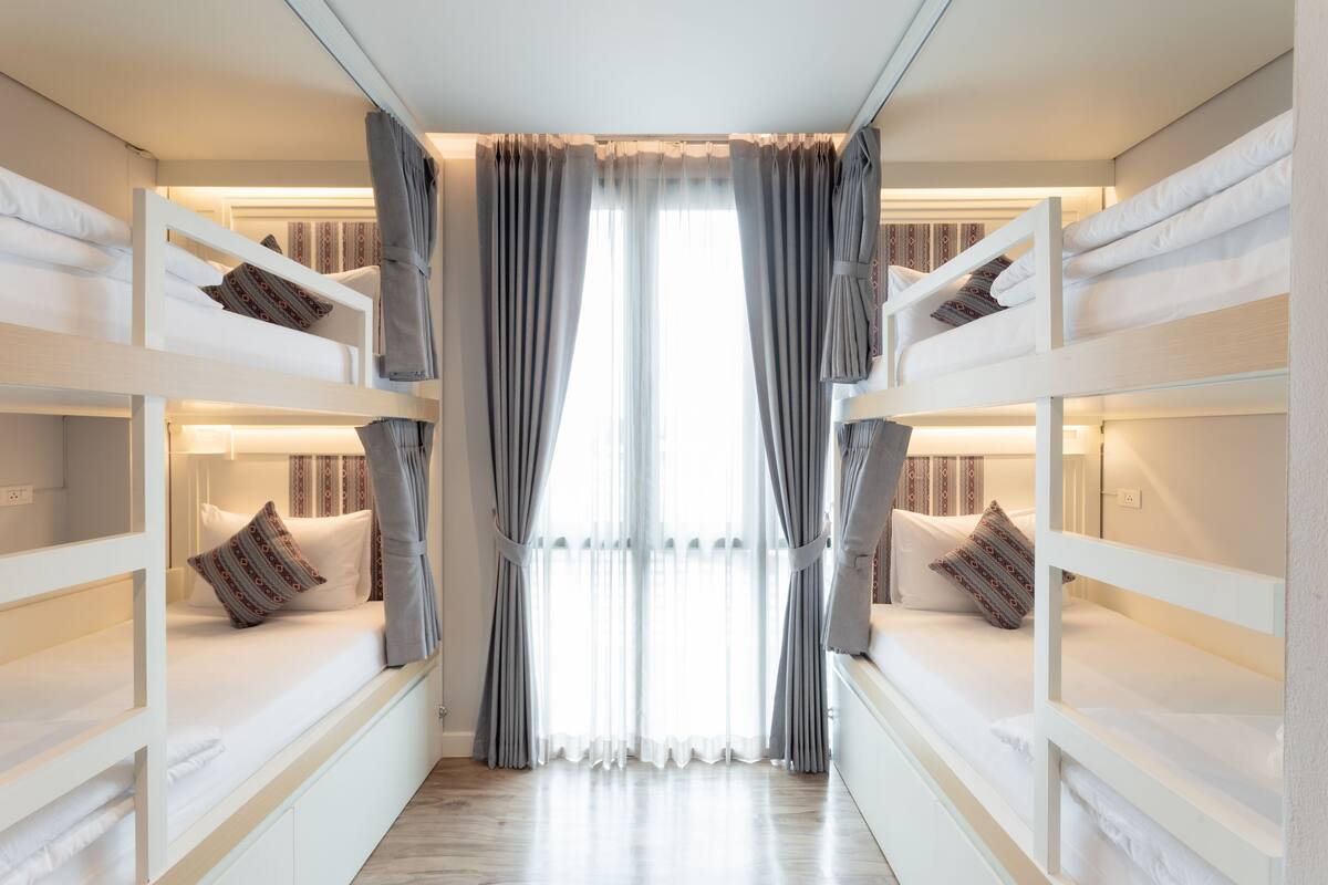 A room with bunk beds, a large window with sheer curtains, and a wooden floor.