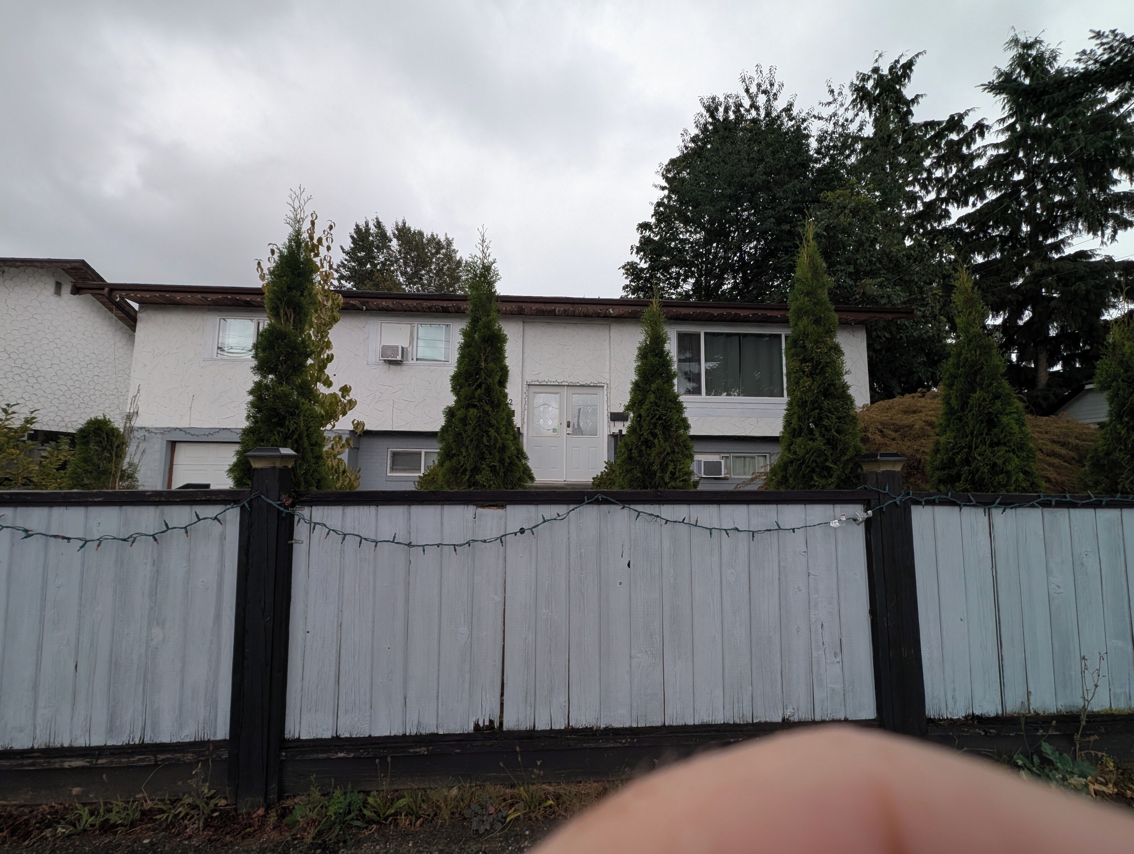 Un quartier résidentiel avec une clôture blanche, une maison avec un toit brun et des arbres à feuilles persistantes.
