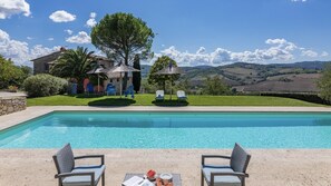 Une piscine rectangulaire aux eaux d’un bleu limpide, entourée d’une terrasse en pierre. On y trouve deux fauteuils de jardin en osier avec des coussins, une table sur laquelle sont disposés des morceaux de pastèque et des verres, ainsi qu’un petit parasol. Au fond, on aperçoit une maison en pierre avec un toit de chaume, un arbre et une vue sur des collines ondoyantes.
