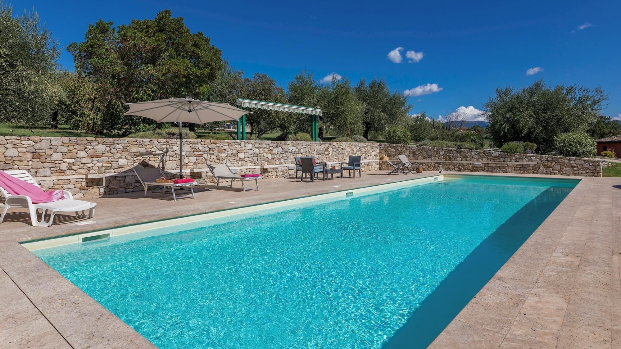 A rectangular swimming pool with clear blue water, surrounded by stone walls and a stone patio. There are several lounge chairs and a covered seating area with a table and chairs.