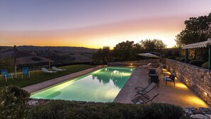 Una piscina rettangolare con muretto in pietra e lettini prendisole, con il tramonto sullo sfondo.