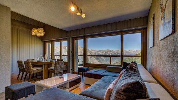 Una sala de estar moderna con un ventanal que ofrece vistas a la montaña, un comedor con mesa y sillas de madera, y un rincón de descanso acogedor con sofá y reposapiés.