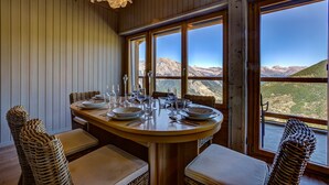 Comedor con mesa de madera para cuatro personas, ventanas amplias con vistas a la montaña, sillas de mimbre y una lámpara colgante.