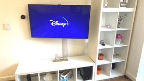 A flat-screen TV mounted on a white entertainment unit with a Disney+ logo, a small shelf with decorative items, and a white cabinet with glassware.