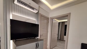 Una habitación moderna con una televisión de pantalla plana sobre un mueble, aire acondicionado instalado en el techo y una puerta que da a otra habitación.